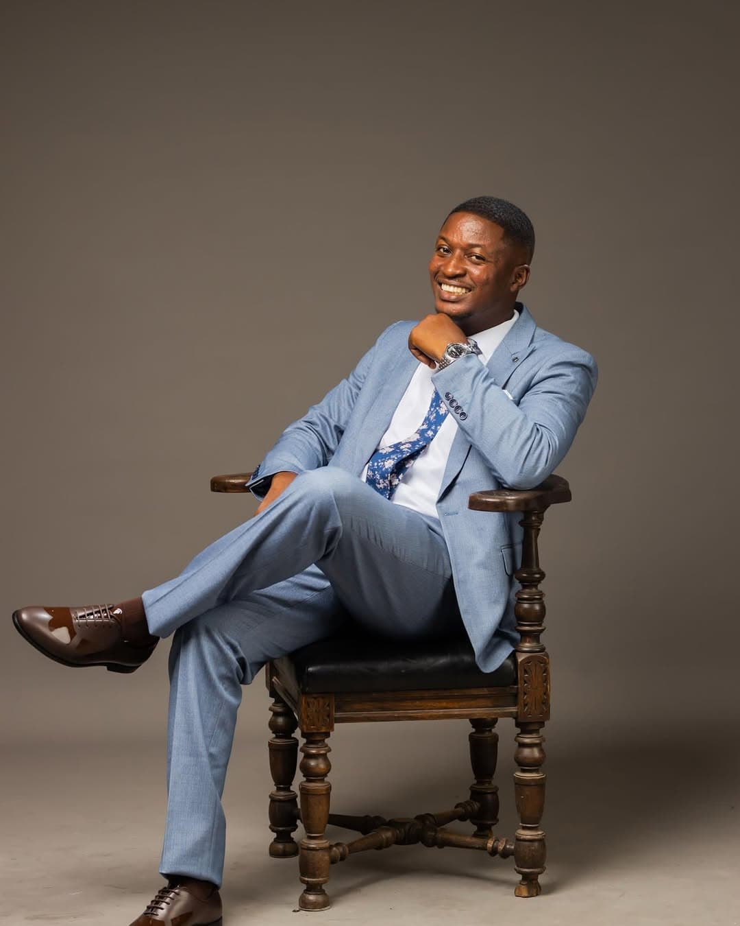 Rev. Theophilus Badger in professional light blue suit sitting in ornate wooden chair, smiling warmly